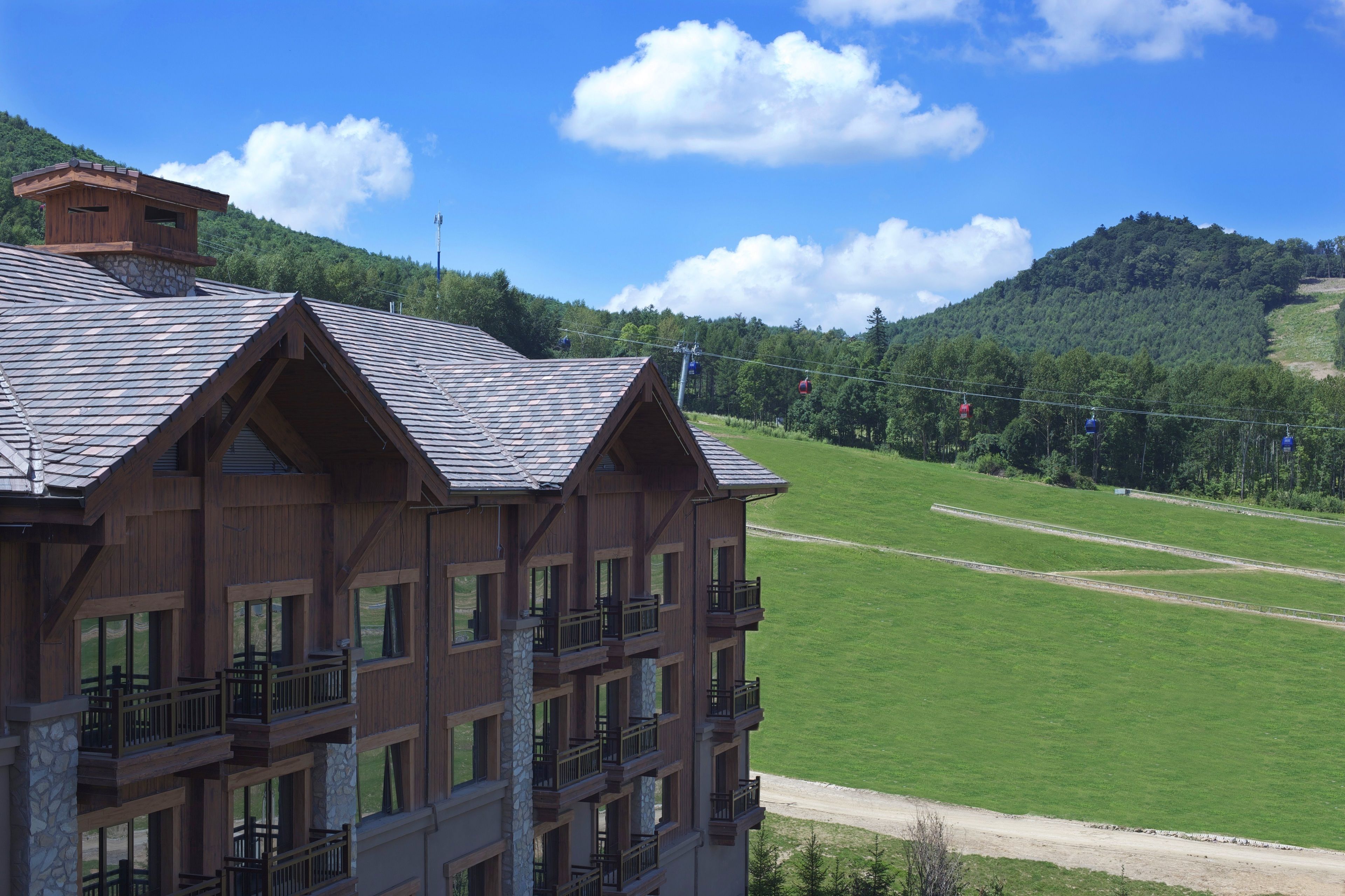 A multi-story wooden building with balconies, set against a backdrop of a green field and forested hills.