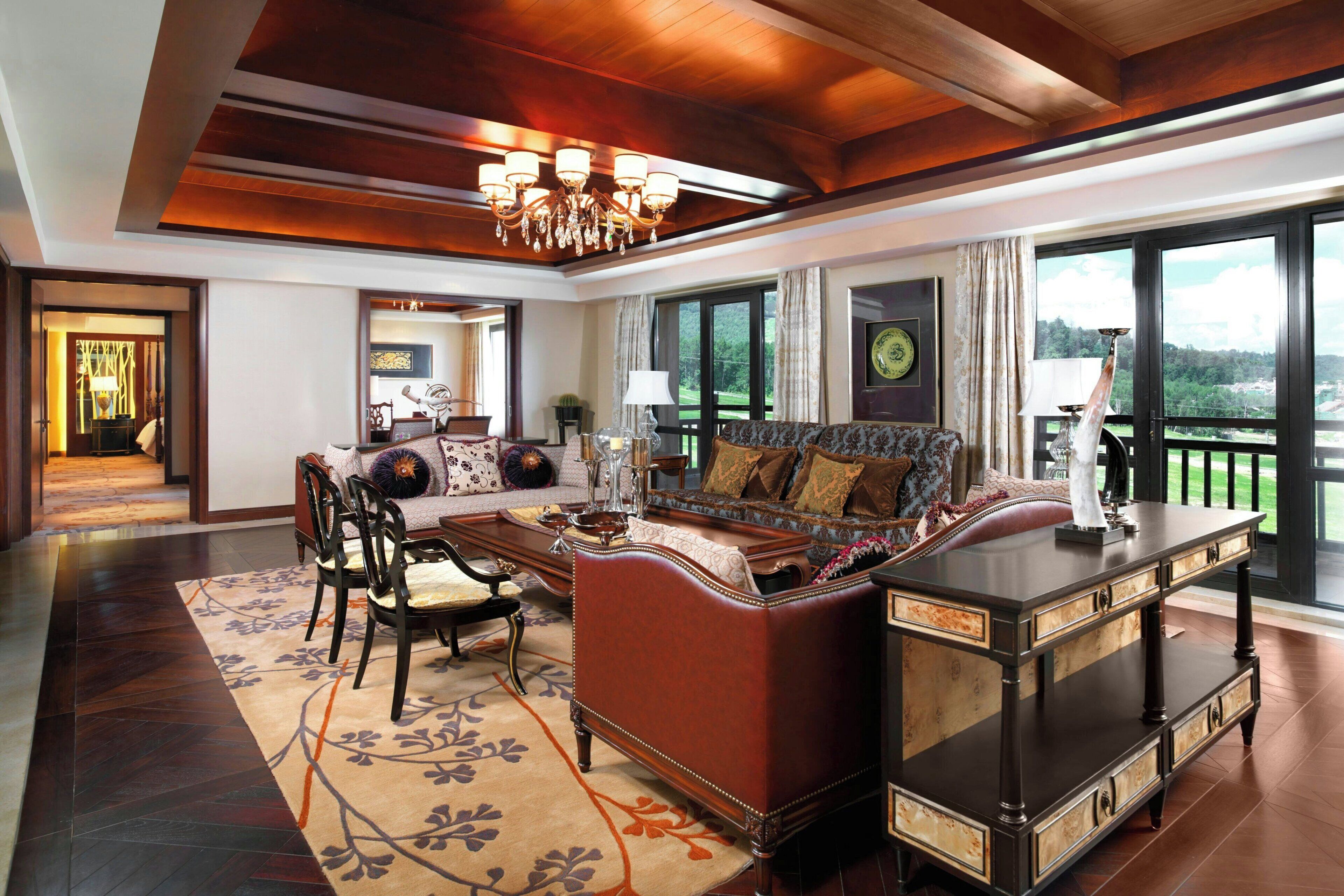 A spacious living room with a large sofa, a coffee table, and a sideboard. There is a chandelier hanging from the ceiling and large windows offering a view of the outdoors.