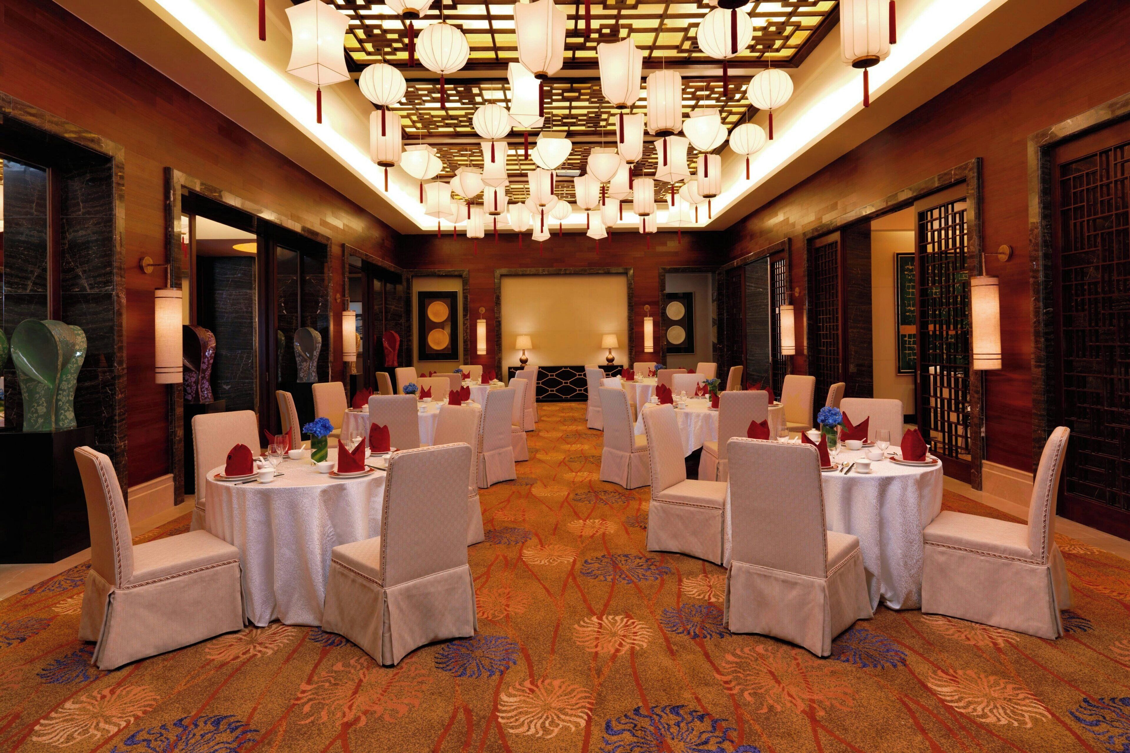 A banquet hall with round tables and chairs, a patterned carpet, and traditional lanterns.