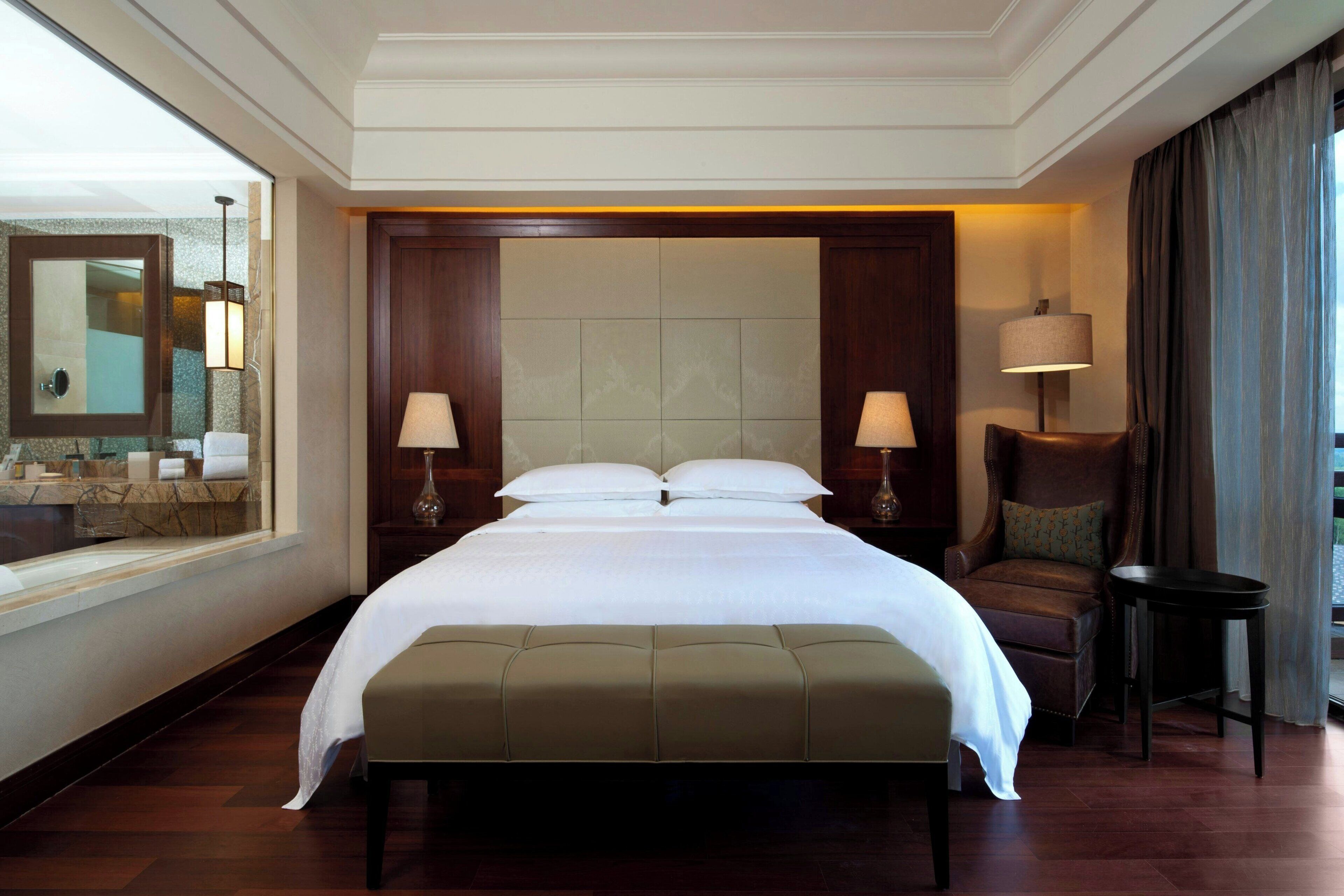 A hotel room with a large bed, two bedside tables with lamps, a chair, a small table, and a large mirror.