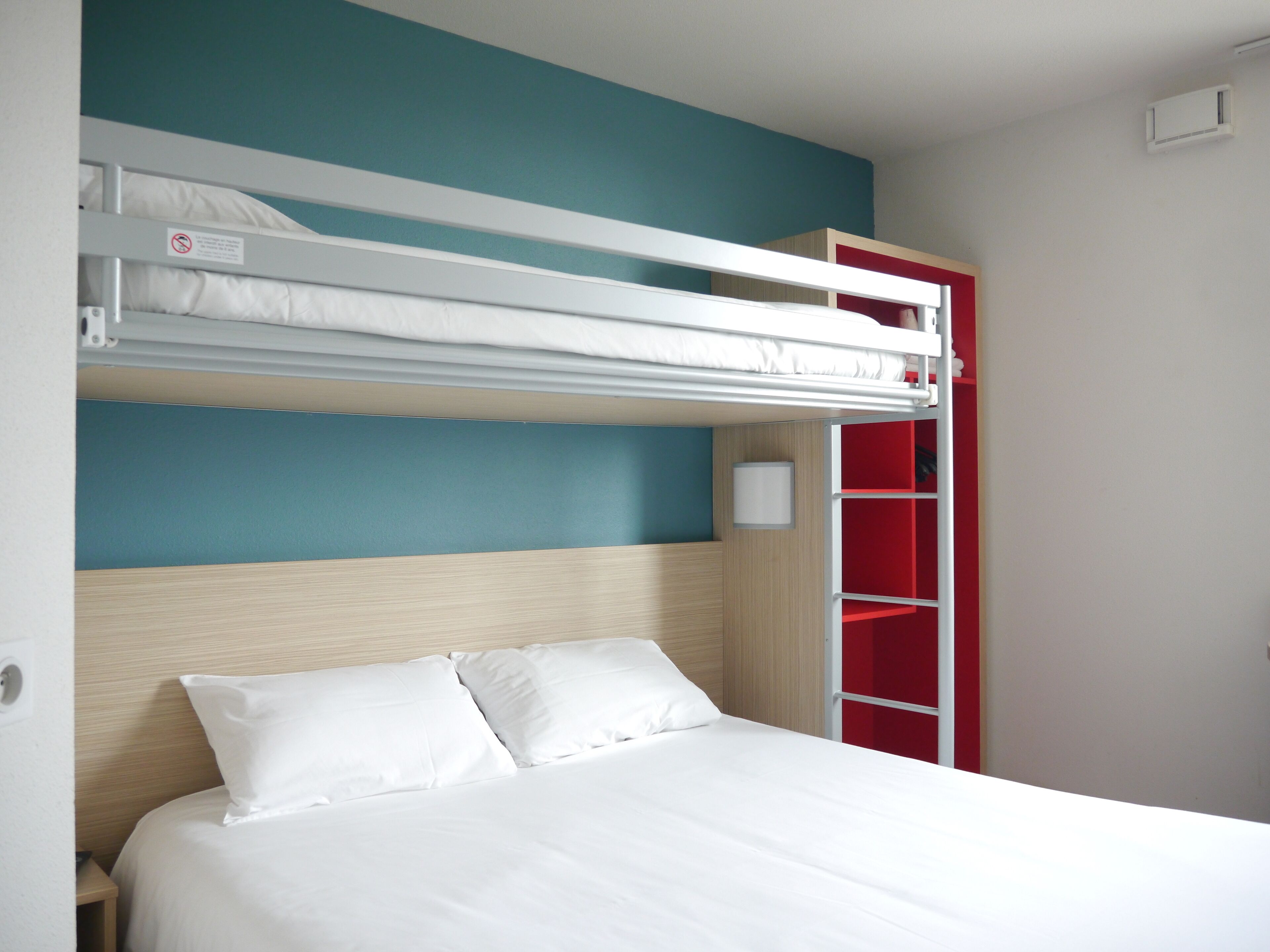 Un lit superposé avec une literie blanche, une tête de lit en bois et un meuble de rangement rouge.