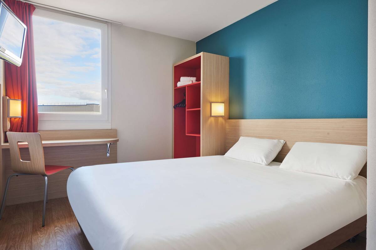 Hotelkamer met een bed, een bureau met stoel, een televisie, een kast en een raam met gordijnen.