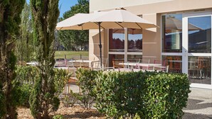 Een terras met tafels en stoelen, een grote parasol, en een gebouw met grote ramen op de achtergrond.