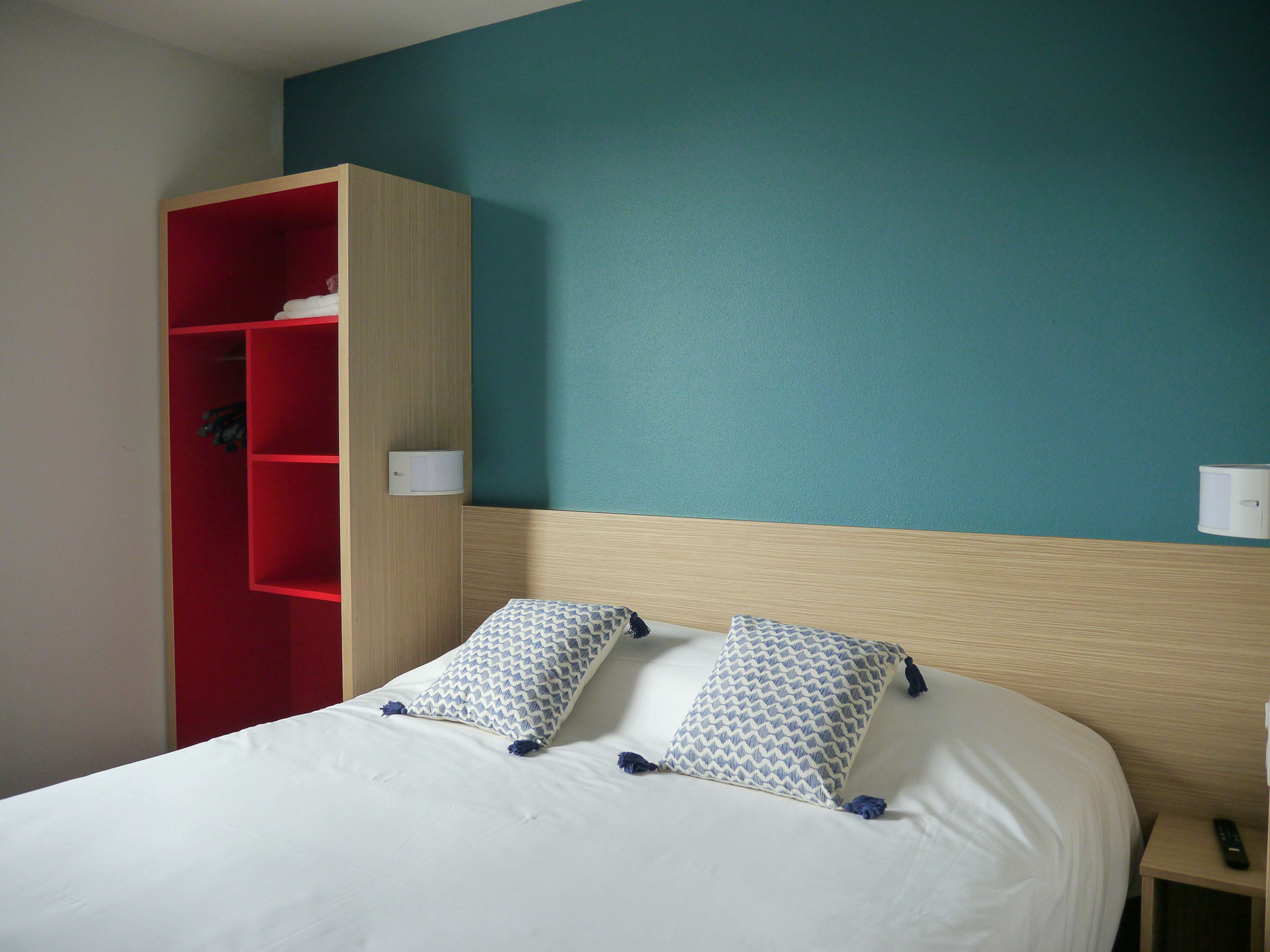 Habitación de hotel con cama, dos almohadas estampadas, un armario rojo y una mesita de noche con control remoto.