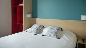 Habitación de hotel con cama, dos almohadas estampadas, un armario rojo y una mesita de noche con control remoto.