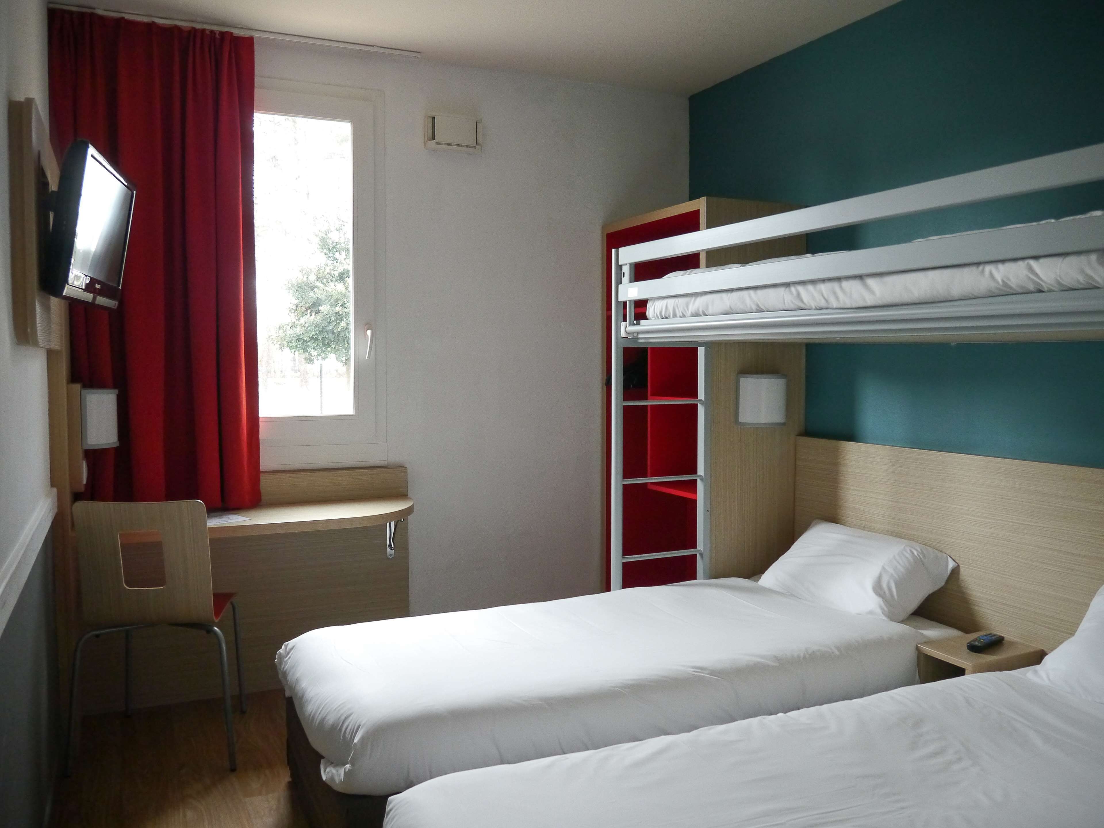 Una habitación de hotel con literas, un escritorio con silla, un televisor y una ventana con cortinas rojas.