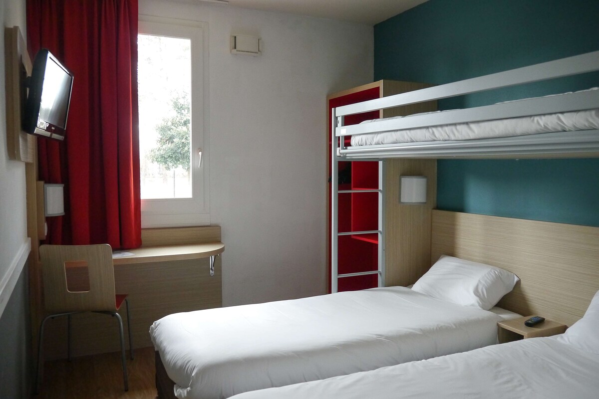 A hotel room with bunk beds, a desk with a chair, a television, and a window with red curtains.