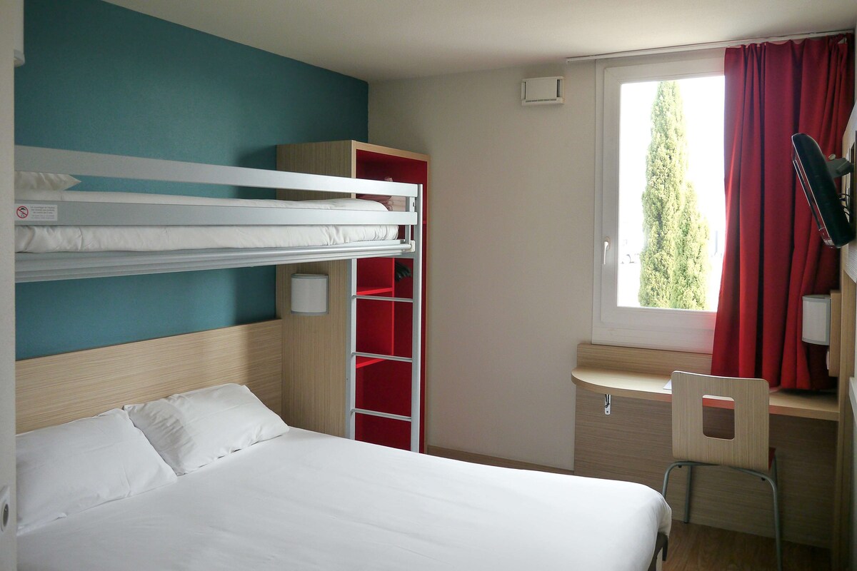 A room with a bunk bed, a desk with a chair, a red wardrobe, and a window with curtains.