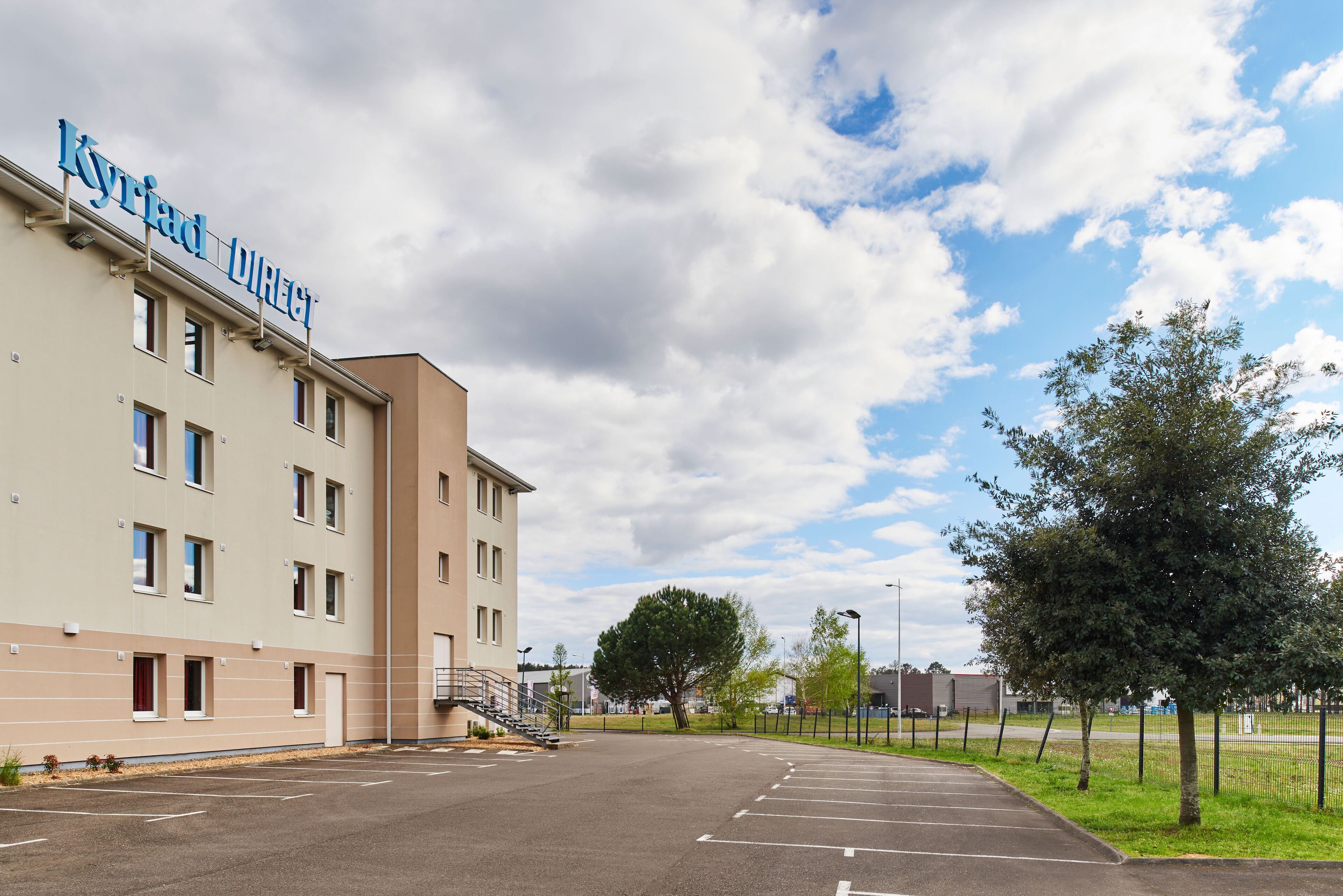 A hotel named 'Kyriad Direct' with a parking lot and a clear sky.