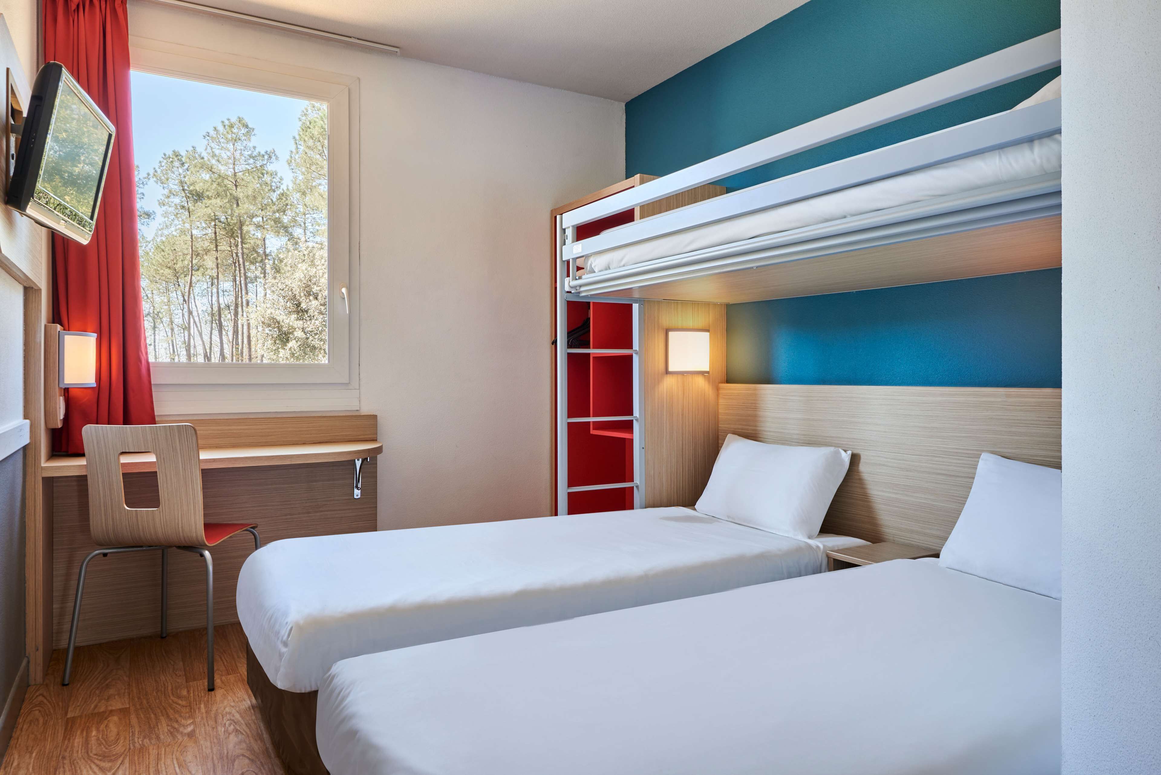 A hotel room with two beds, a desk, a chair, a TV, and a window with a view of trees.