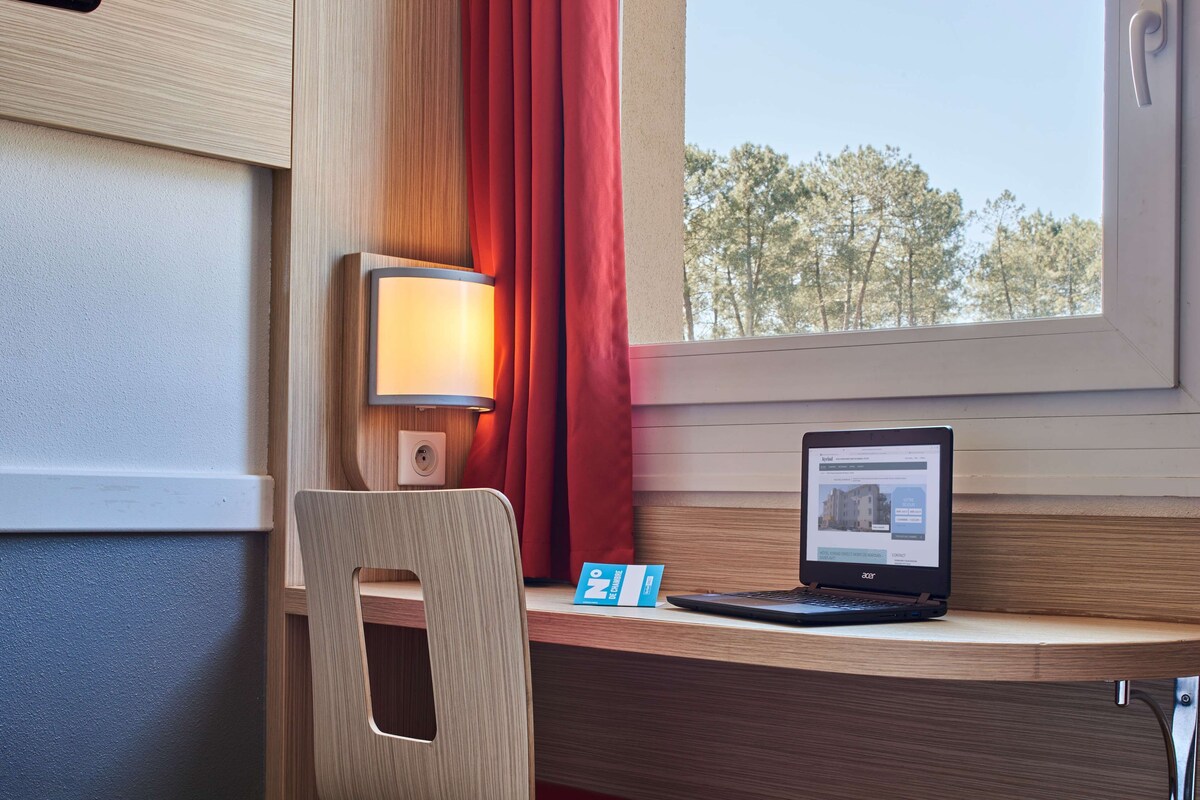 A wooden desk with a laptop, a lamp, and a window with red curtains.