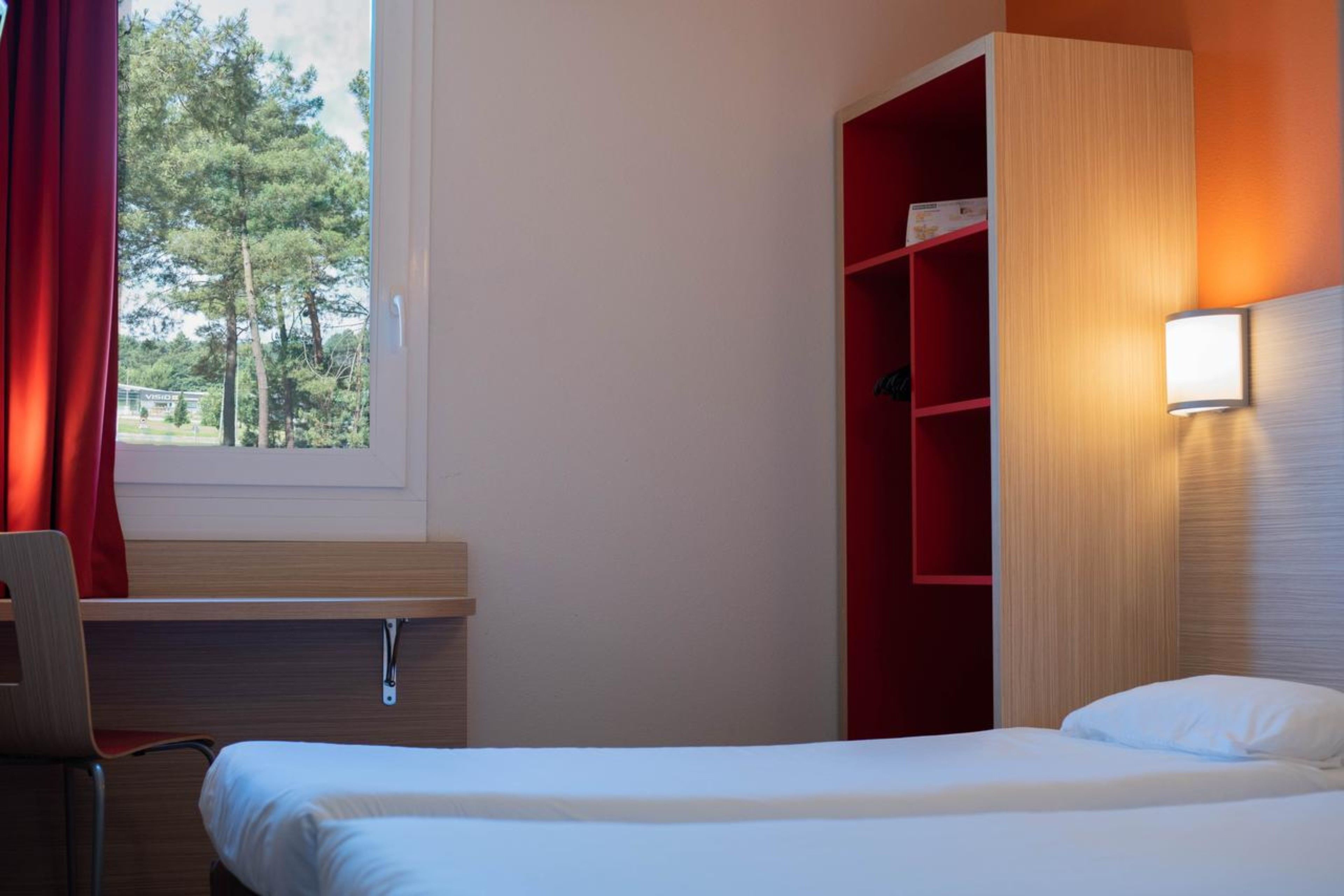 Ein Hotelzimmer mit Bett, Schreibtisch, Stuhl, einem roten Regal und einem Fenster mit Vorhängen.