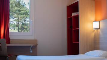 Ein Hotelzimmer mit Bett, Schreibtisch, Stuhl, einem roten Regal und einem Fenster mit Vorhängen.