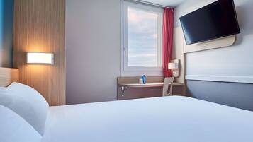 A hotel room with a bed, a desk with a chair, a TV mounted on the wall, and a window with a view of the sky.