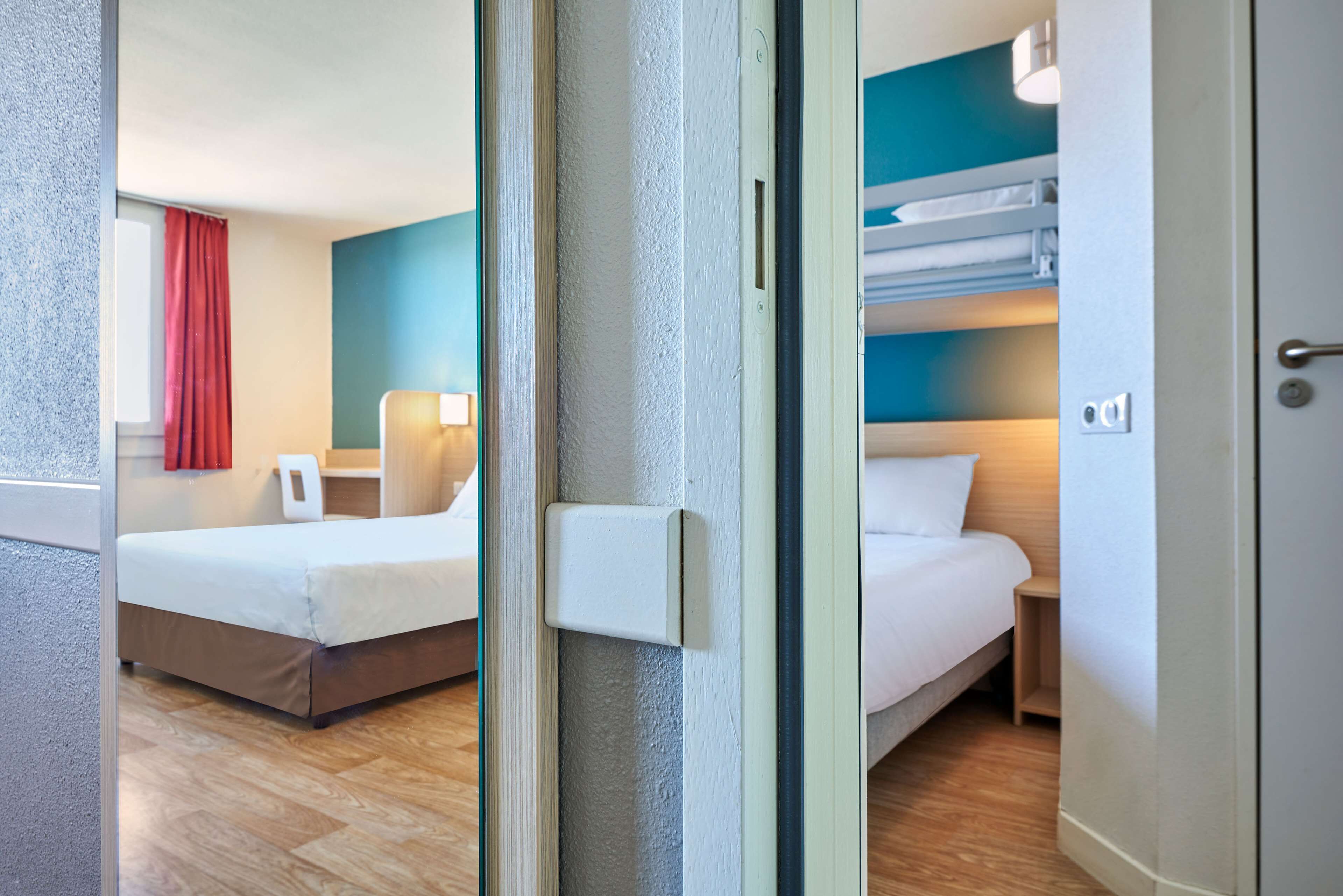 A hotel room with a bed, a bedside table, a lamp, and a window with a red curtain.