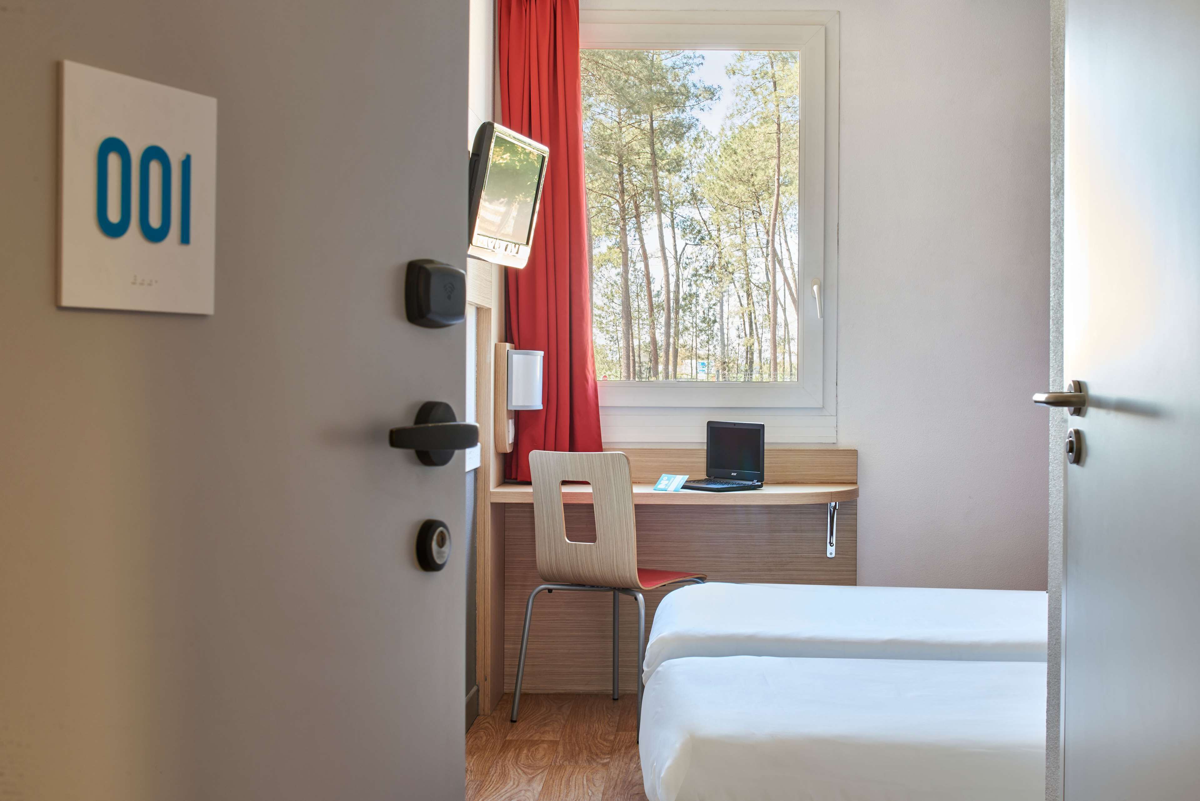Een hotelkamer met een bed, een bureau met een laptop, een stoel, een raam met uitzicht op bomen en een deur.