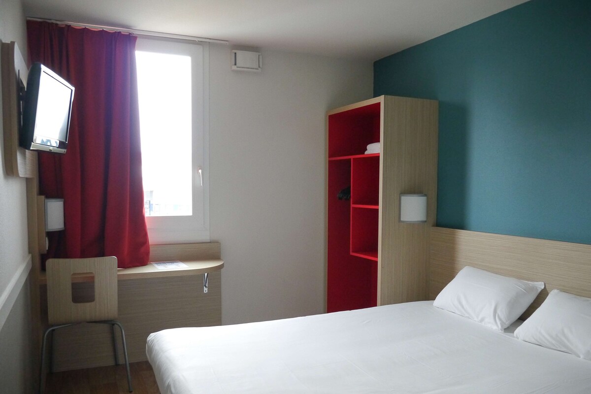 A hotel room with a bed, a desk, a chair, a TV, a wardrobe, and a window with curtains.