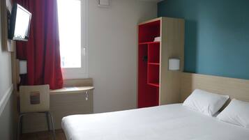 Habitación de hotel con cama, escritorio, silla, televisión, armario y ventana con cortinas.
