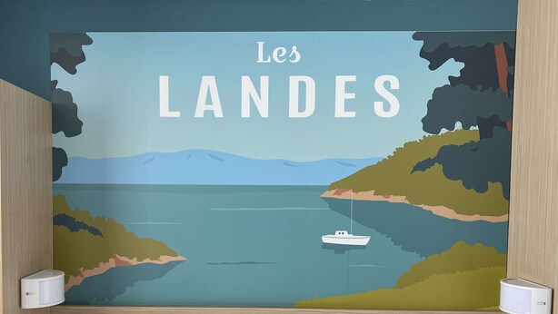Een slaapkamer met een muurschildering van “Les LANDS” dat een kustlandschap met een boot toont.
