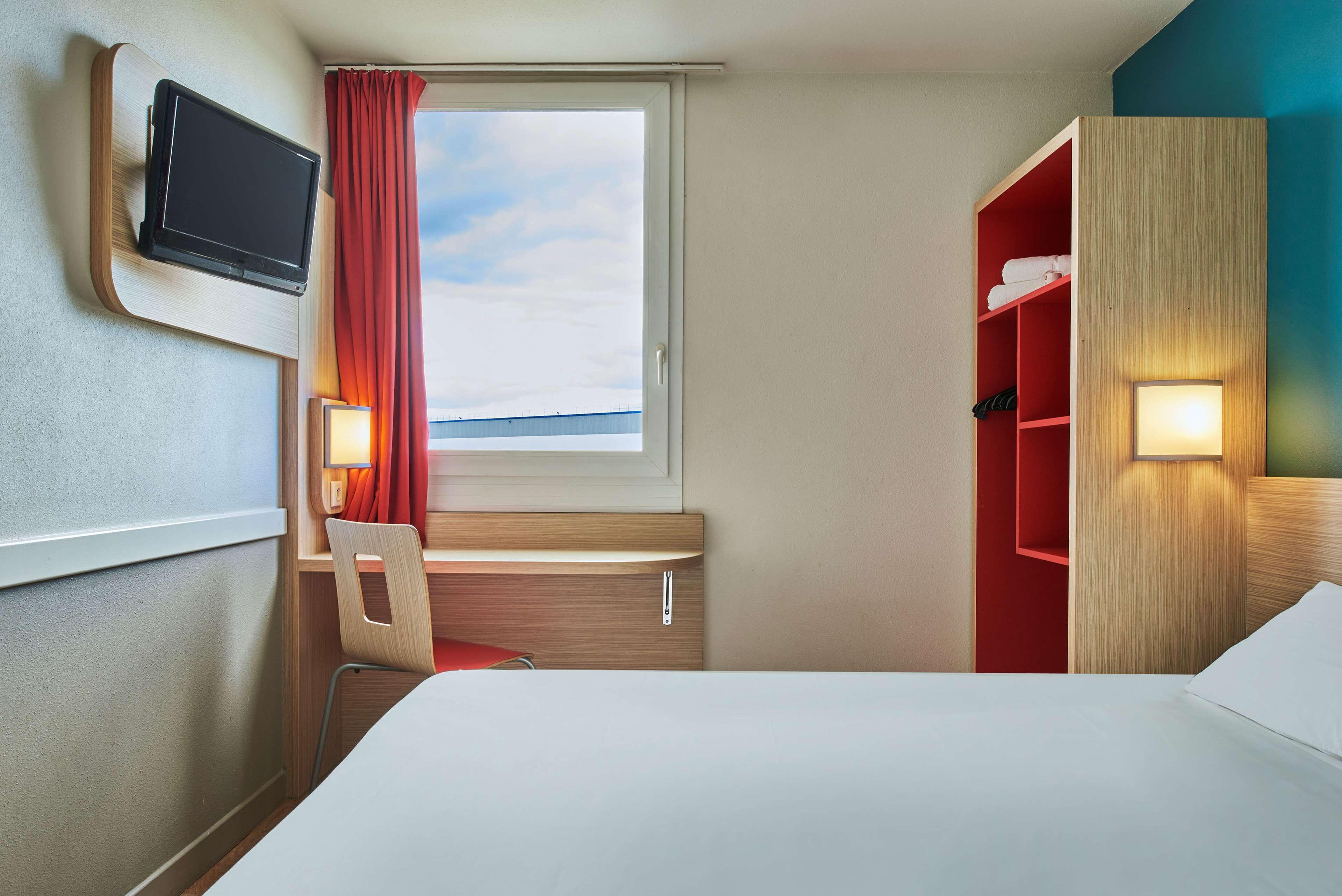 A hotel room with a bed, a desk with a chair, a TV mounted on the wall, a window with curtains, and a wardrobe with shelves.