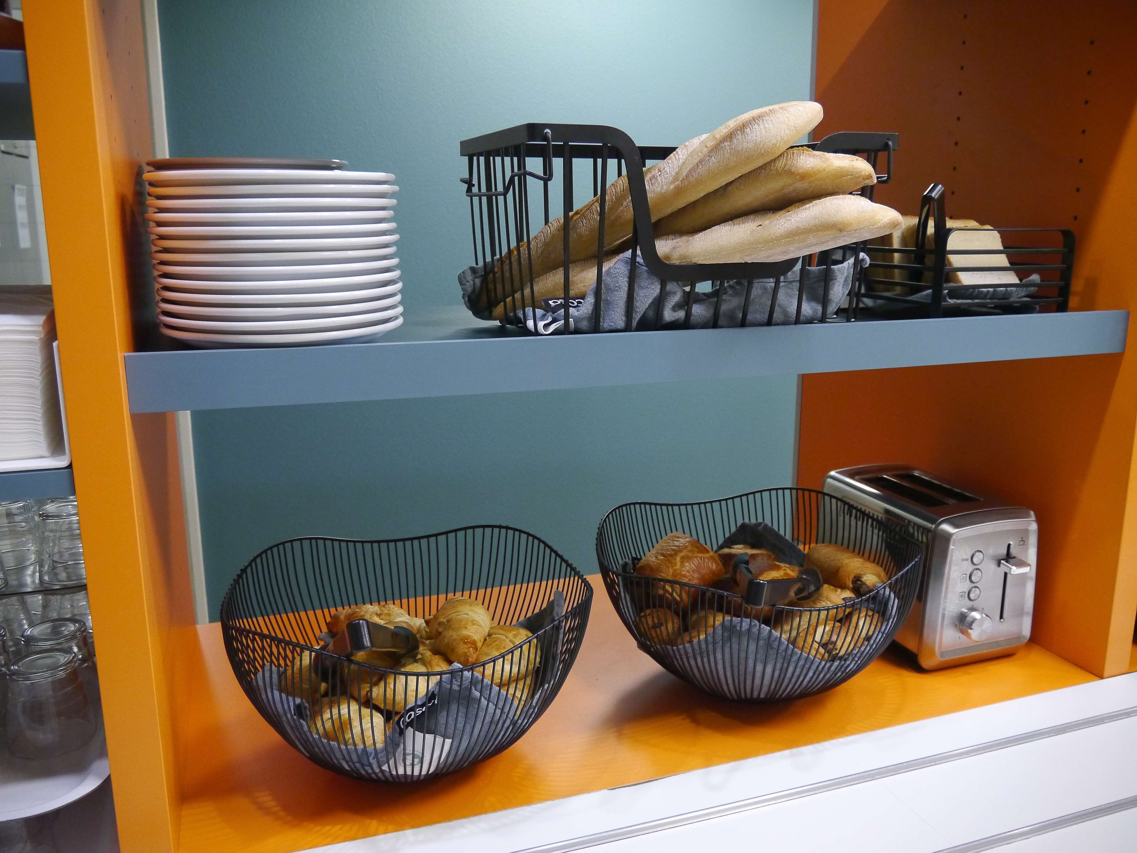 A kitchen counter with two wire baskets containing baked goods, a stack of white plates, and a toaster.