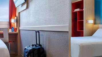 Habitación de hotel con cama, escritorio y silla, televisor montado en la pared, maleta en el suelo y un mueble de almacenamiento rojo con luz.
