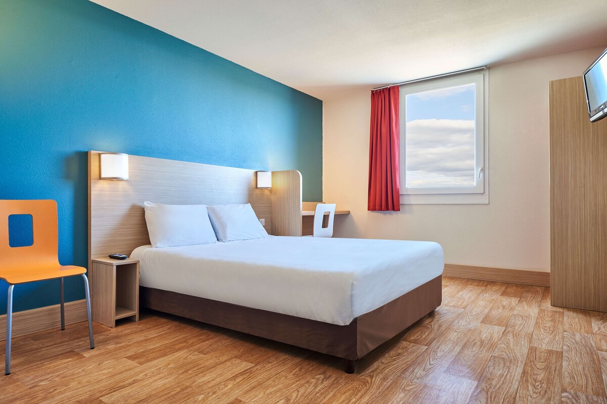 A neatly arranged hotel room with a bed, a chair, a nightstand, and a TV mounted on the wall.