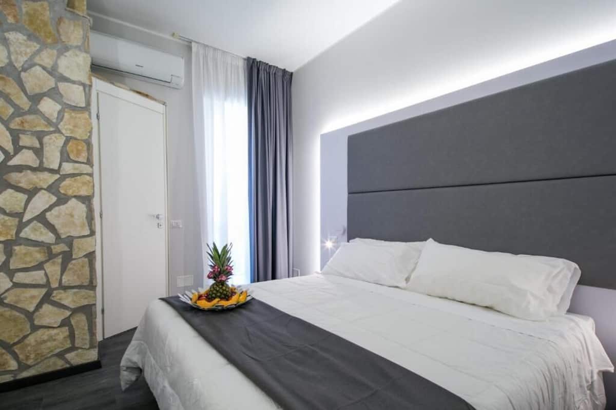 A hotel room with a stone accent wall, a bed with white linens, and a fruit bowl on the bed.