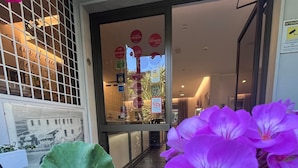 Entrada de un hotel con puerta de cristal, vista del interior y una planta con flores moradas en primer plano.