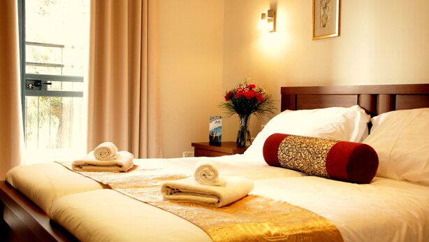 A neatly made bed with two rolled towels, a wooden headboard, and a nightstand with a vase of flowers. - City Center Jerusalem (Jerusalem)