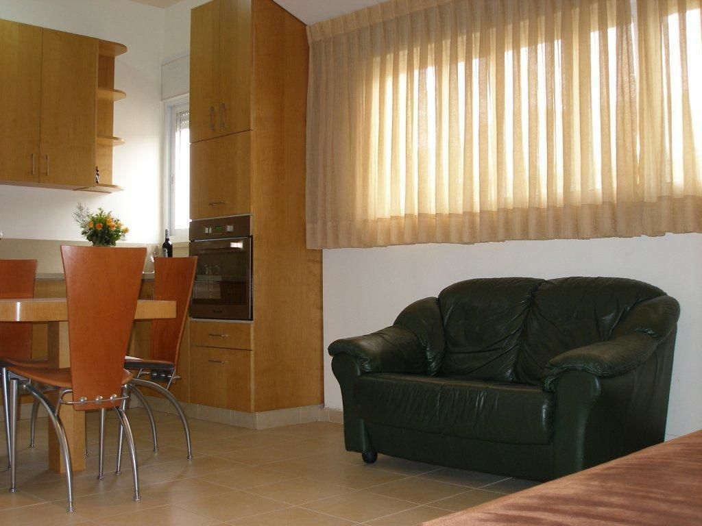 Een keuken met houten kasten, een eettafel met stoelen, een groene leren bank en een raam met dunne gordijnen.