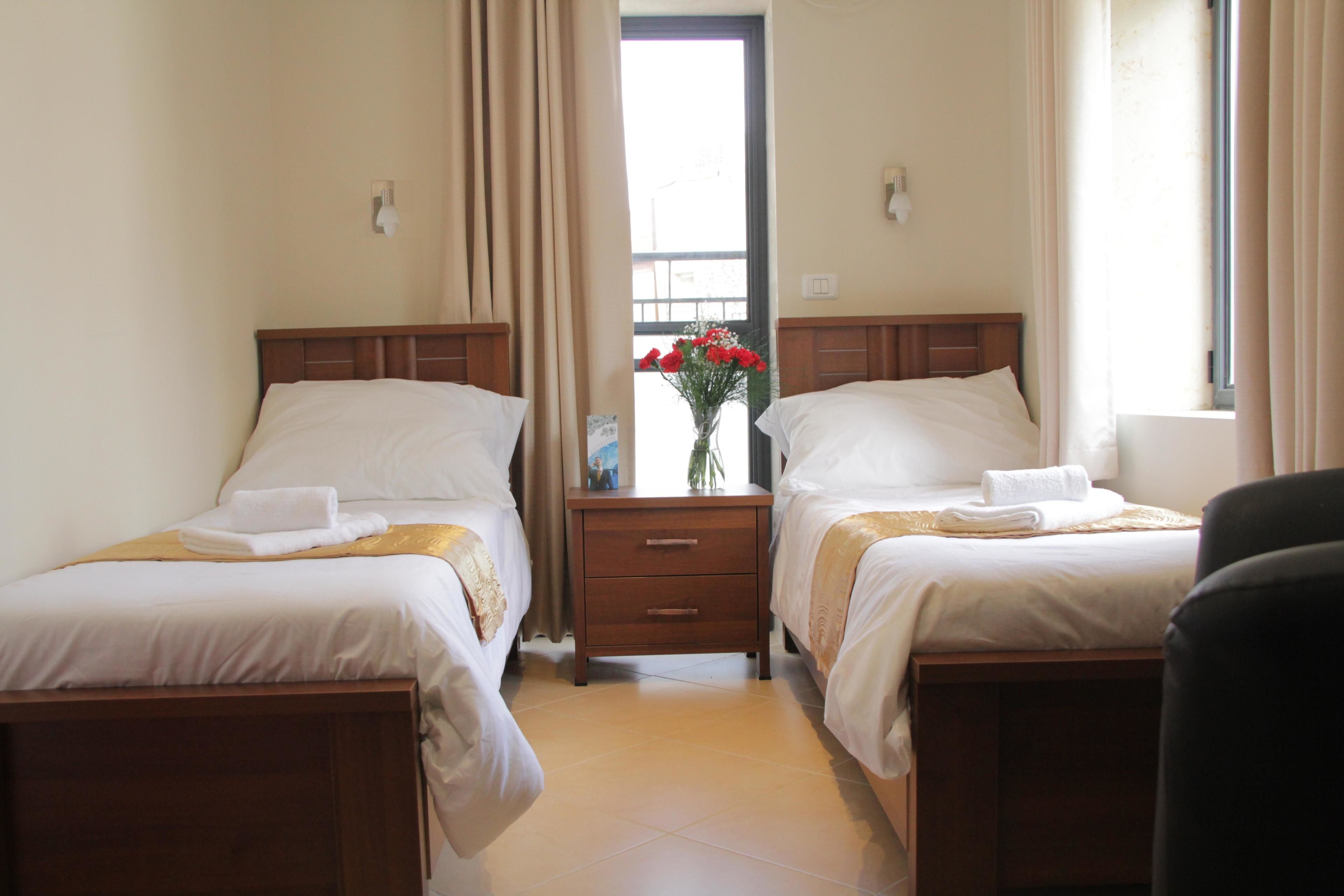 Ein Hotelzimmer mit zwei Einzelbetten, einem Holfnachtkästchen, einer Blumenvase und einem Fenster mit Vorhängen.