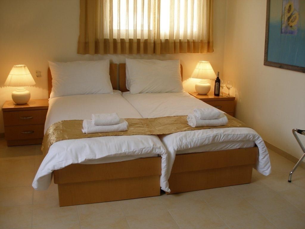 A double bed with white linens, two bedside tables with lamps, a wooden bench at the foot of the bed, and a wheelchair in the corner.
