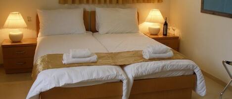 A double bed with white linens, two bedside tables with lamps, a wooden bench at the foot of the bed, and a wheelchair in the corner.