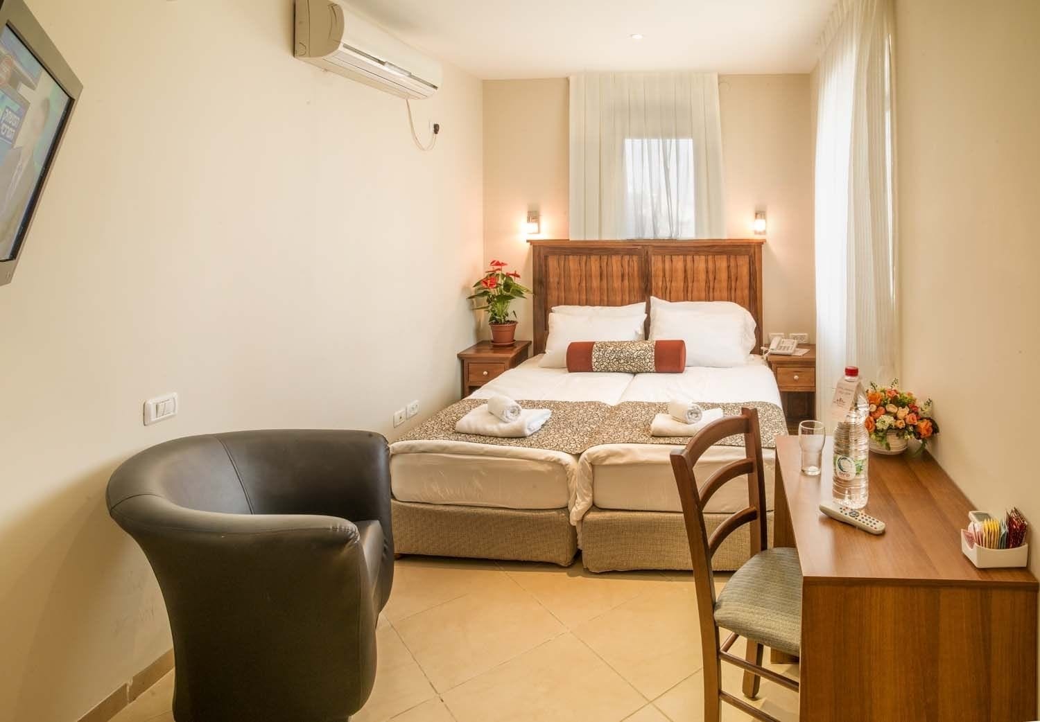 Una habitación de hotel con cama, una silla, un escritorio con una botella y vasos, y una planta en maceta.