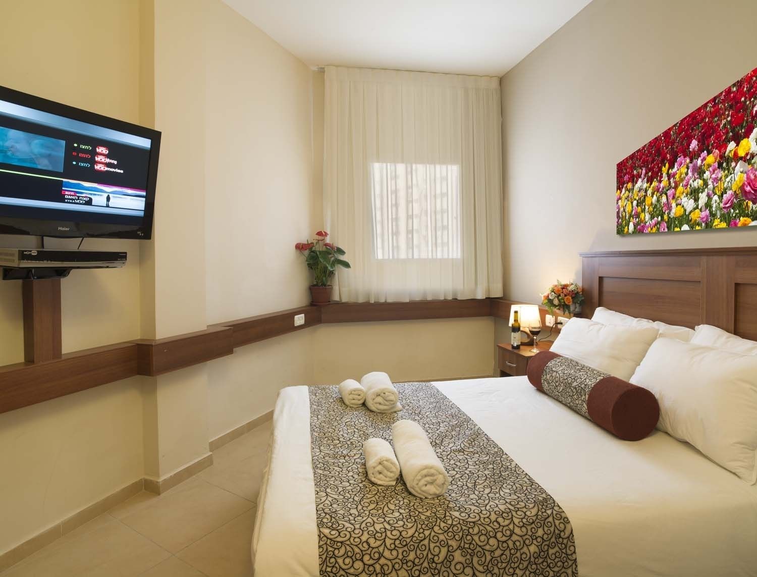 A hotel room with a bed, a TV, a window with curtains, a painting, and a nightstand with a lamp and flowers.