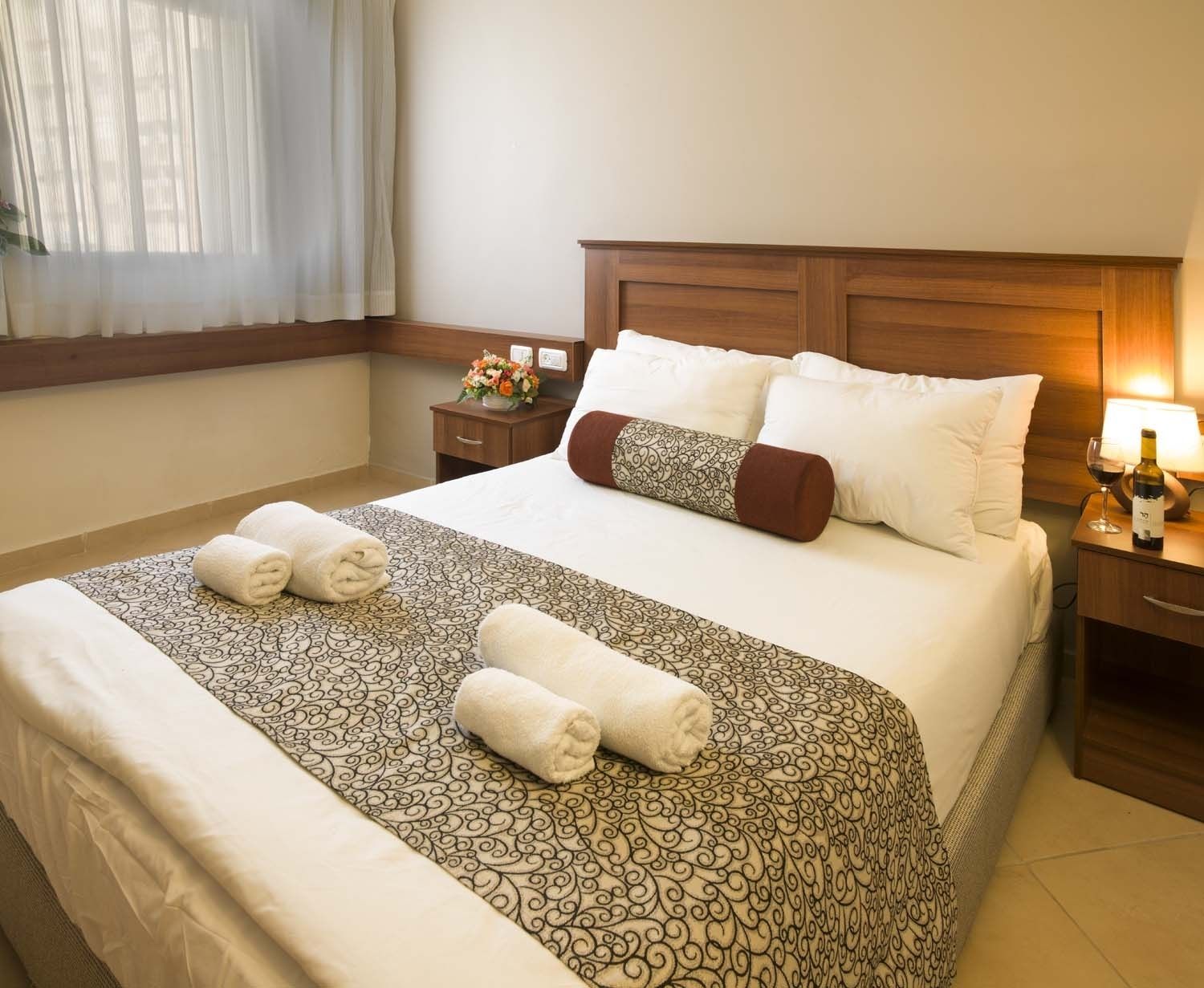 Una cama bien hecha con una colcha estampada, dos toallas enrolladas y una mesita de noche con una lámpara y una botella de vino.