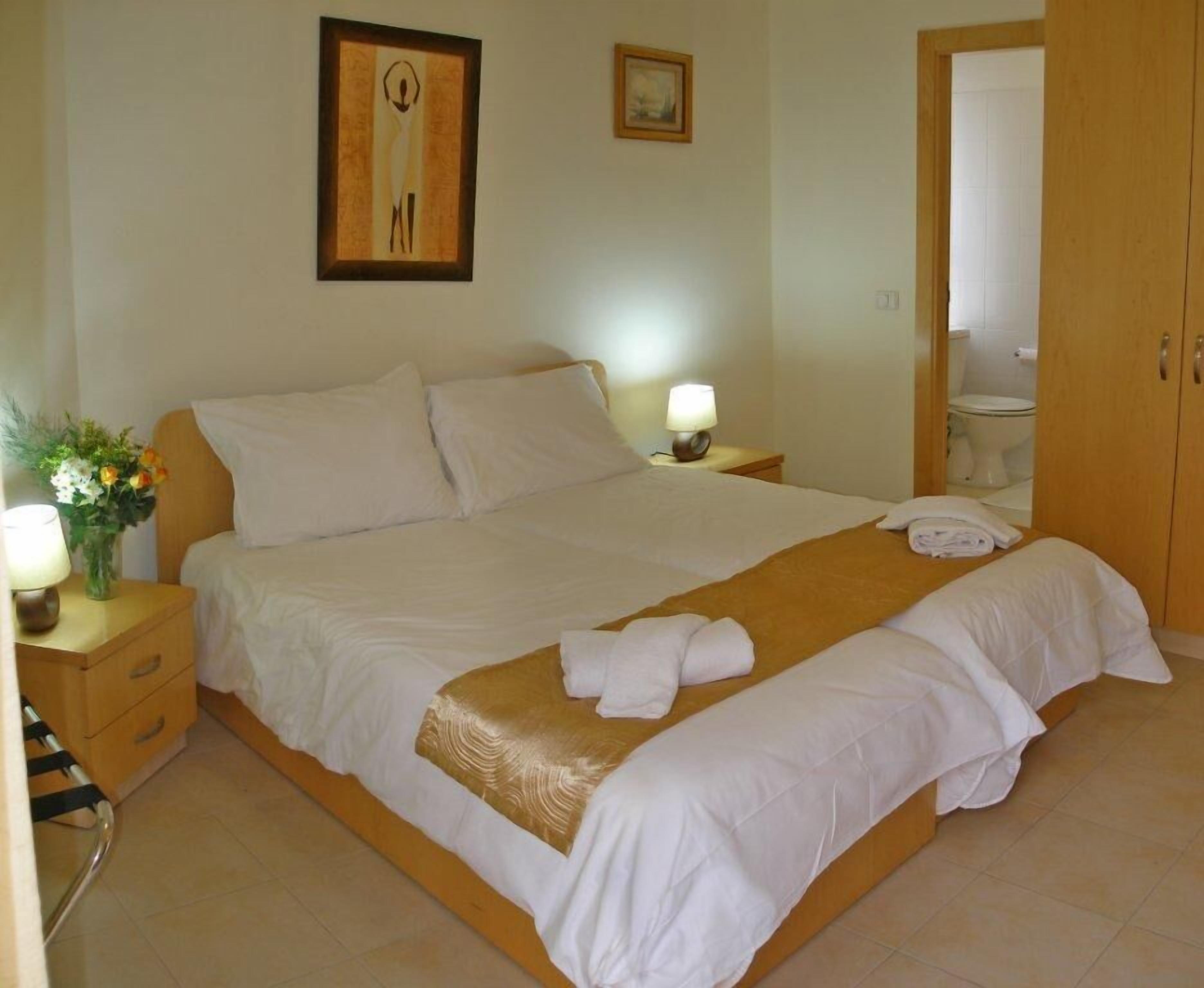 A neatly made bed with white linens and a gold accent, a bedside lamp, and a vase with flowers on a wooden nightstand.