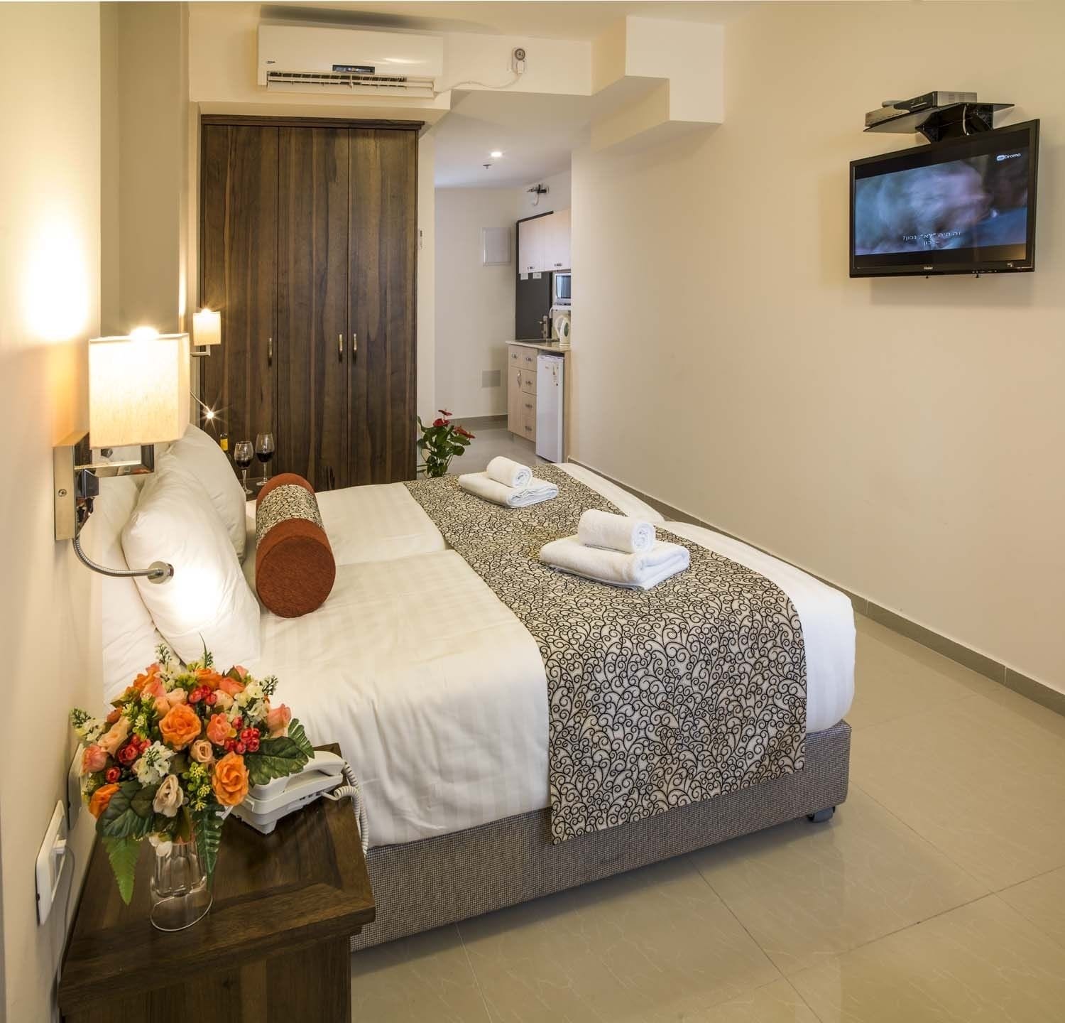 Una camera d'albergo con un letto, una TV, una scrivania con una lampada, un vaso di fiori e un armadio.