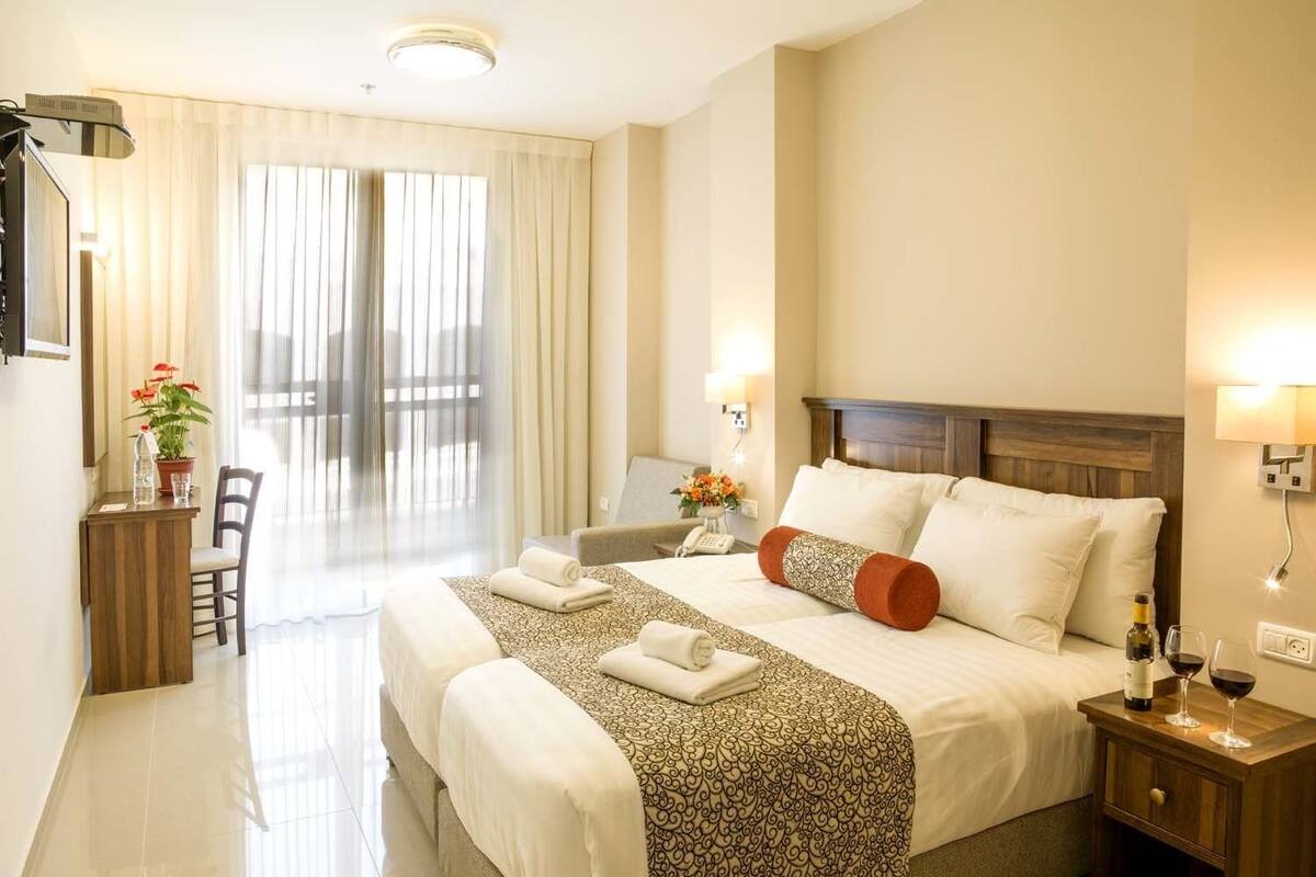 Une chambre d’hôtel avec un grand lit, un bureau avec une chaise, une petite table avec une bouteille et des verres, et une vue sur l’extérieur par la fenêtre.