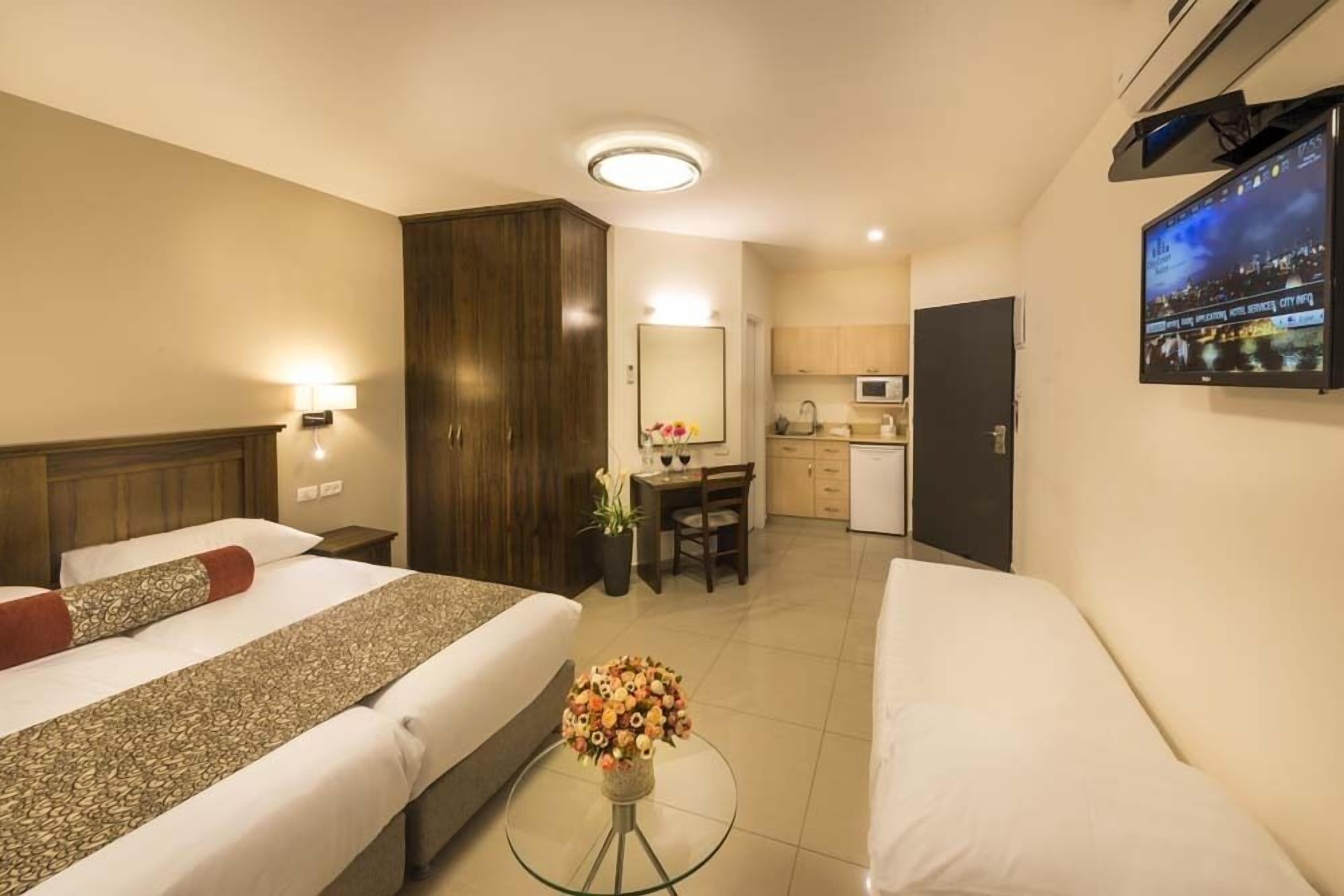 Habitación de hotel con dos camas, un escritorio, una silla, una mesita con un florero, un televisor en la pared y una kitchenette al fondo.