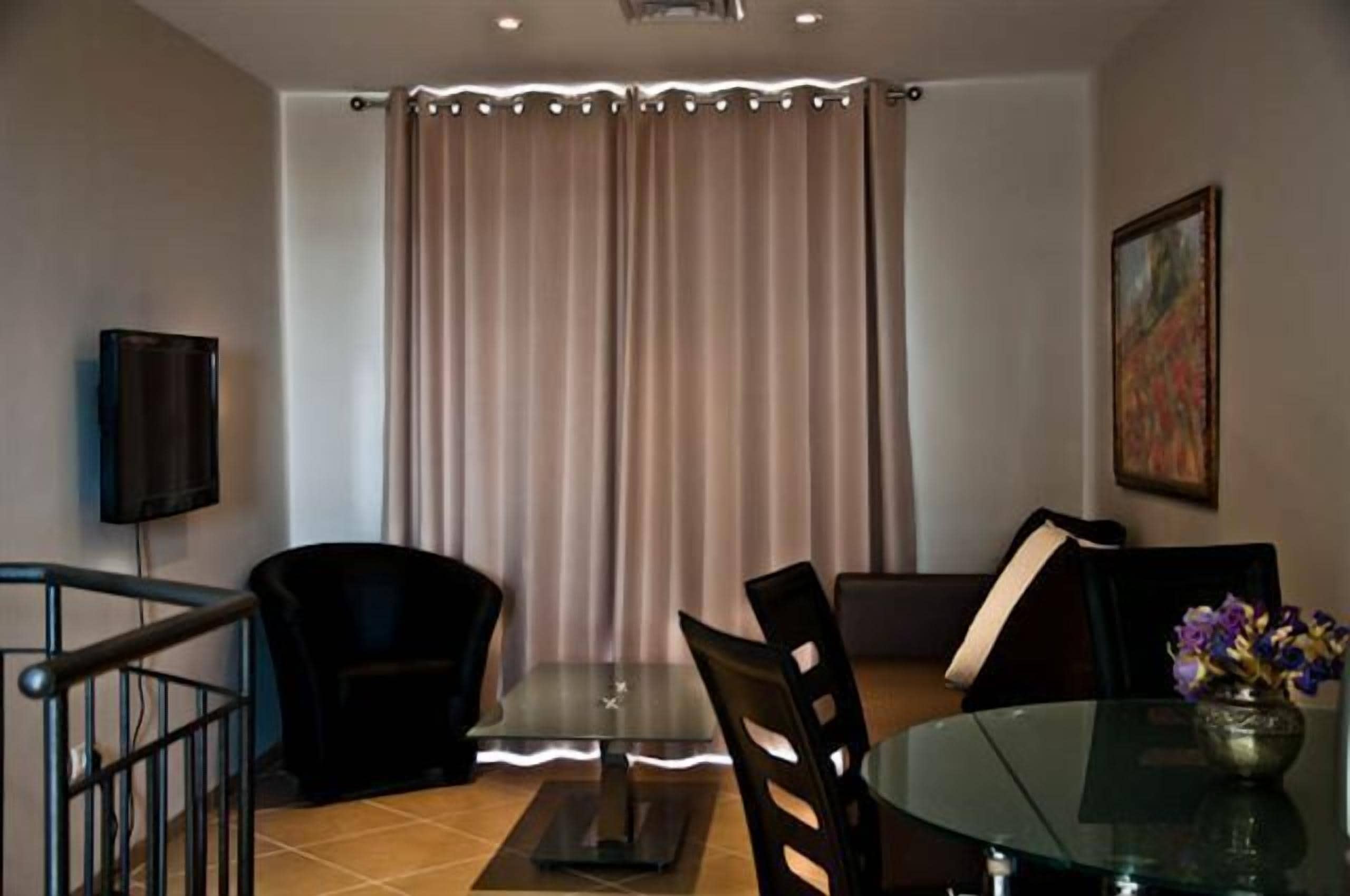 A dining area with a glass table, black chairs, a TV on the wall, and a curtain.