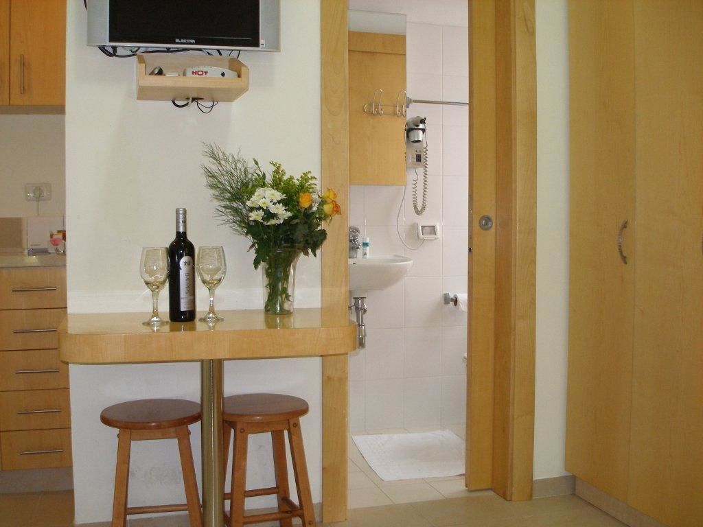 A small kitchen with a wooden table, two stools, a wine bottle, and a vase with flowers.