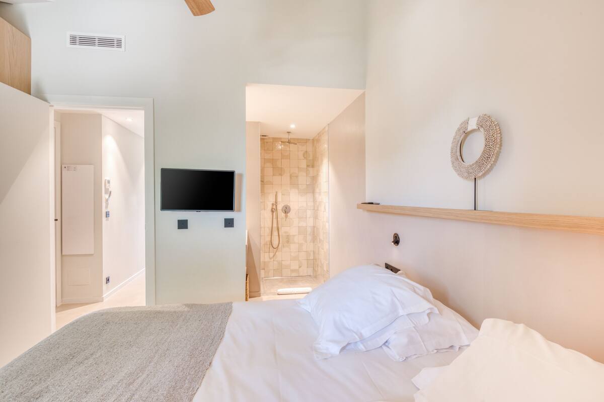 Une chambre à coucher comprenant un lit, un téléviseur fixé au mur, un espace douche et une porte donnant sur une autre pièce.