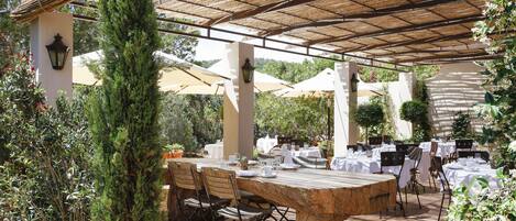 Un espace extérieur aménagé pour les repas, avec des tables et des chaises, entouré de plantes et de parasols.