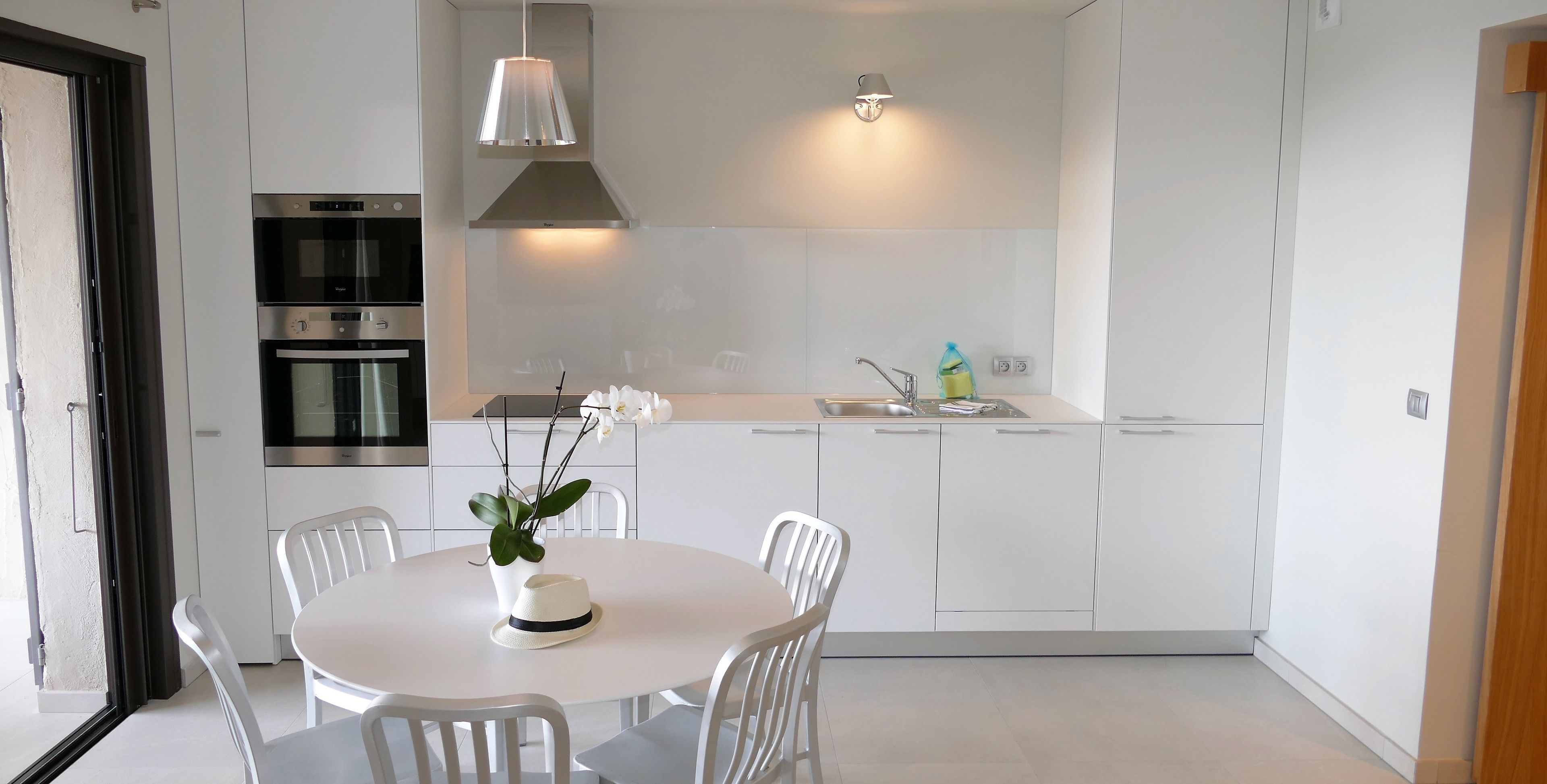 Una cocina moderna con armarios blancos, horno empotrado, lavamanos y comedor con mesa redonda y sillas.
