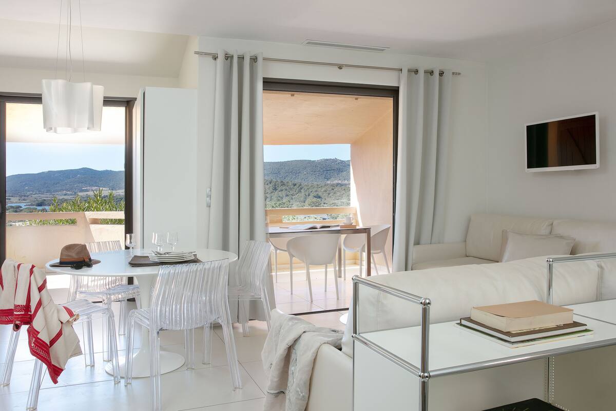 Une pièce moderne et bien éclairée, dotée d’un balcon, d’un coin repas avec des chaises et une table, et d’un canapé sur lequel repose un livre.