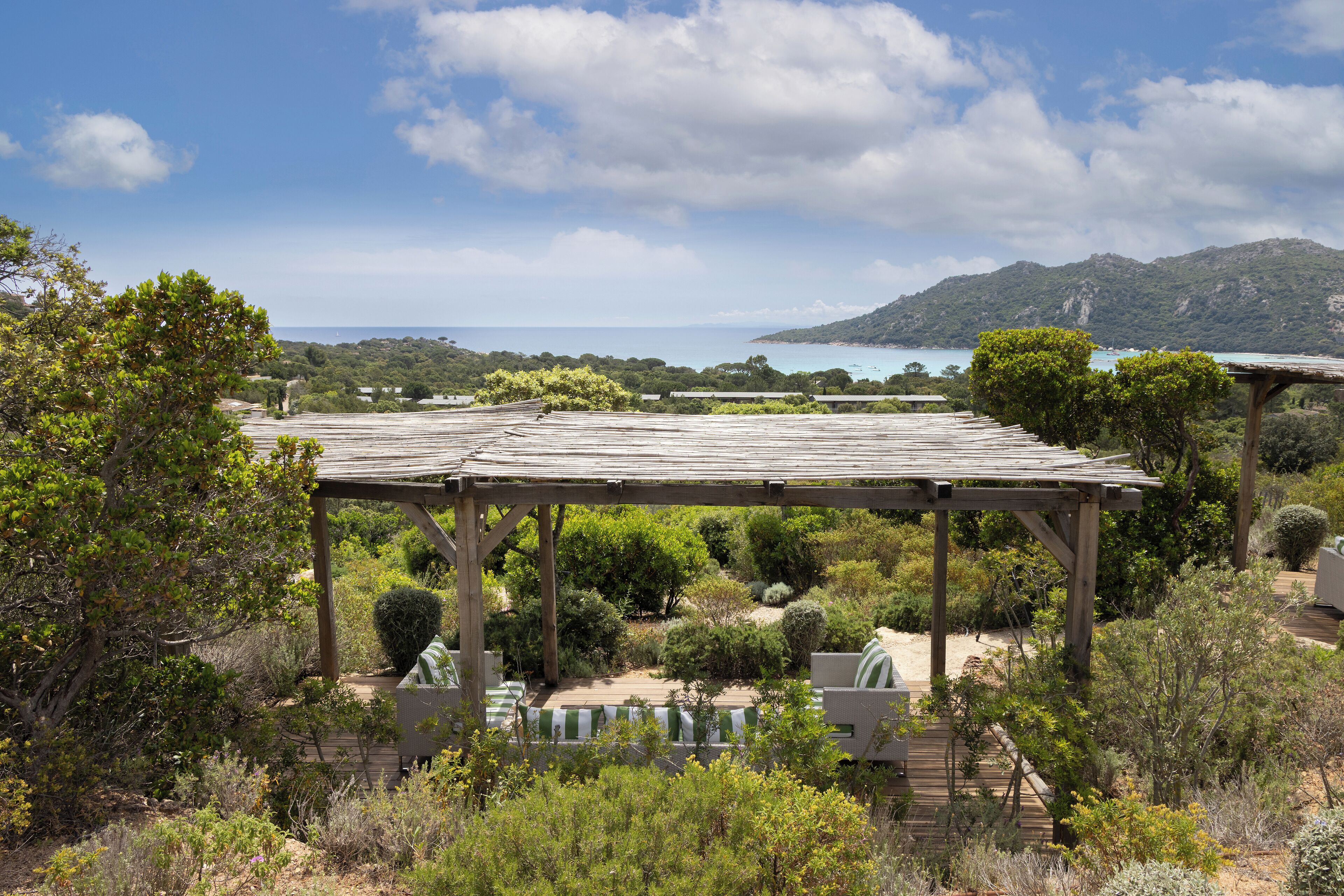Uma pérgula de madeira com área de estar ao ar livre, rodeada por vegetação exuberante e com vista para a paisagem costeira.