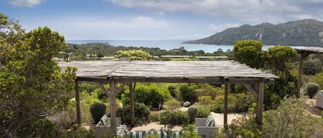 A wooden pergola with outdoor seating, surrounded by lush greenery and overlooking a coastal landscape.