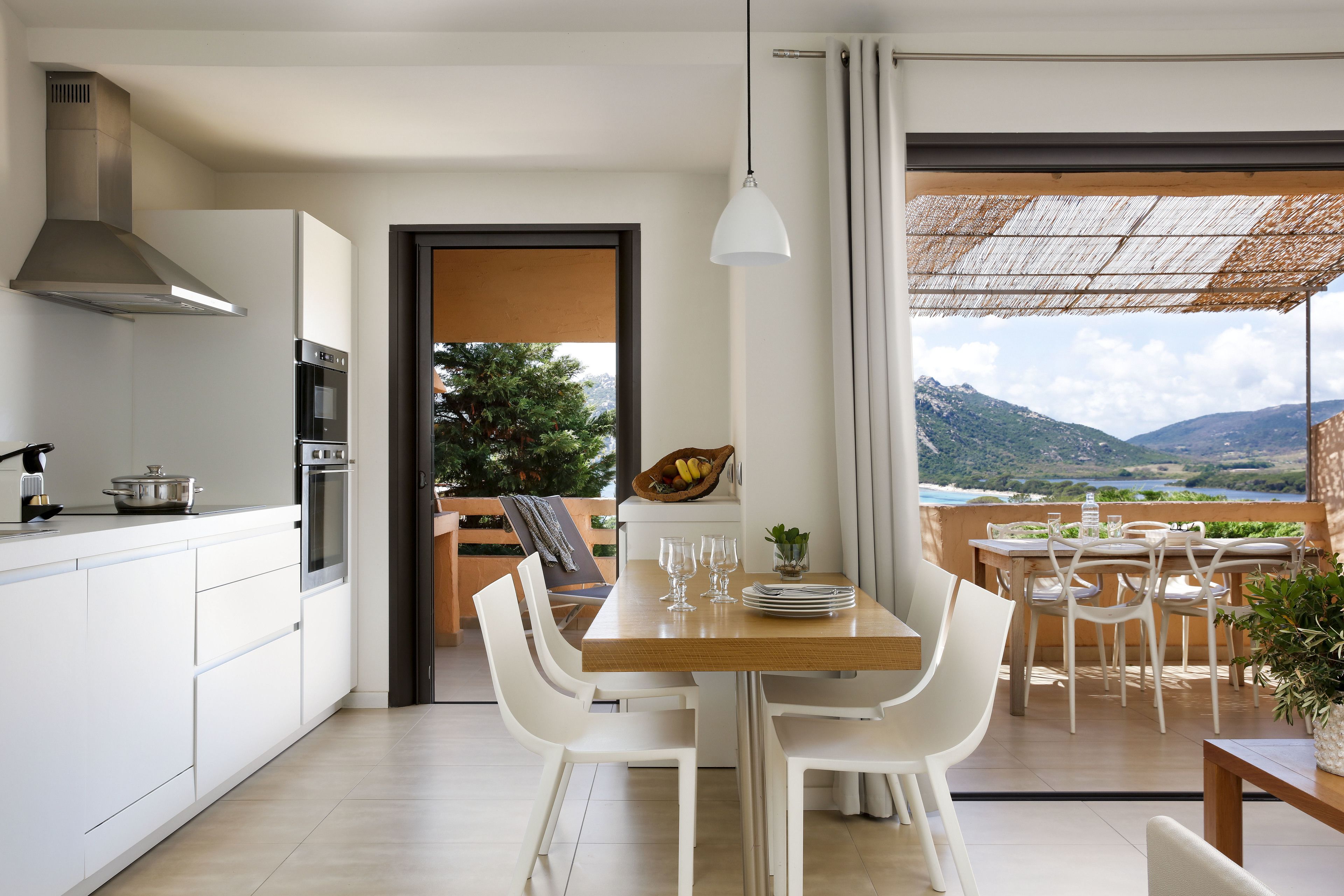 A modern kitchen with white cabinets and stainless steel appliances, a dining area with white chairs and a wooden table, and a view of a mountainous landscape through large windows.